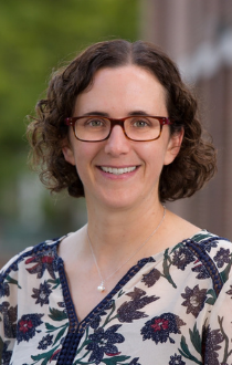 Image: Headshot of Anne McNeil, wearing a floral shirt, short brown hair, with rectangular glasses.