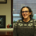 Anne McNeil, white women with curly brown hair, wearing glasses, and a black, grey, and white knitted sweater with flowers and hearts.