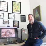 Nate Sanders, white man sitting on desk infront of an ant photo on a desktop monitor. Wearing a blue zip up hoodie.